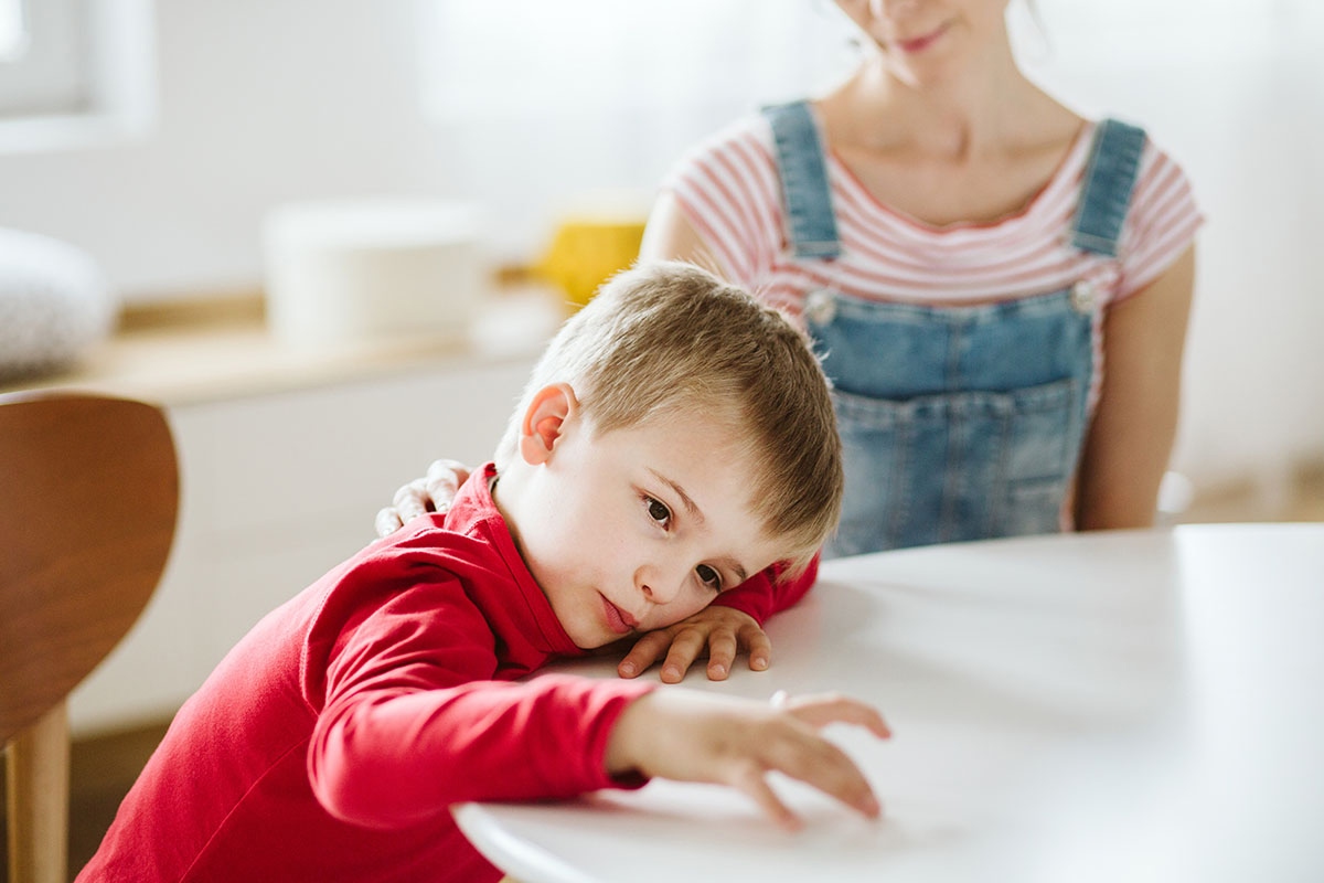 Young child appearing distracted or unfocused while sitting at a table, with a caregiver nearby observing.