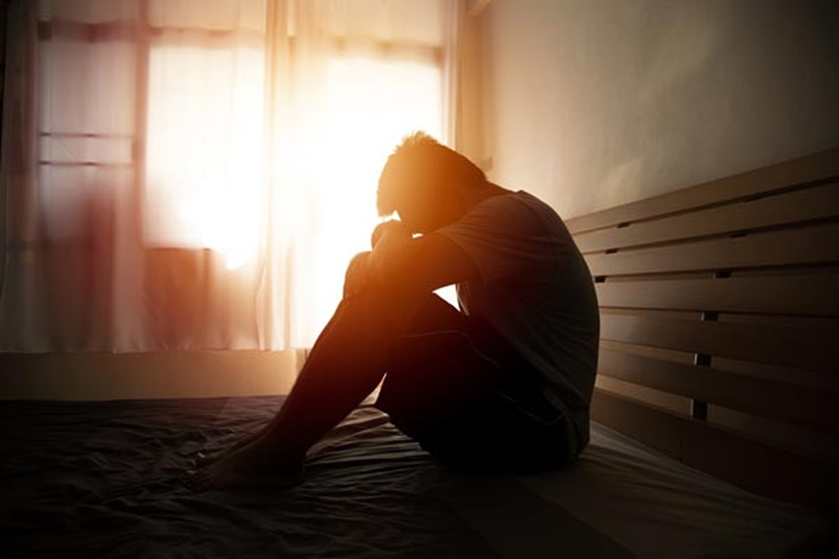 Person sitting alone on a bed with head down, appearing fatigued or distressed, with sunlight coming through a window.